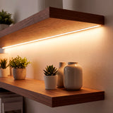 Iluminación lineal continua de tono cálido oculta bajo baldas de madera en muro residencial contemporáneo.