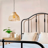 Lámpara de rattan Brisa sobre mesa de noche en dormitorio con cama de forja negra.