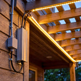Montaje vertical de caja estanca en pared exterior alimentando tiras LED de luz cálida decorativa bajo pérgola.