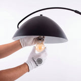 Técnico instalando bombilla de filamento en pantalla semiesférica metálica negra con guantes de protección.