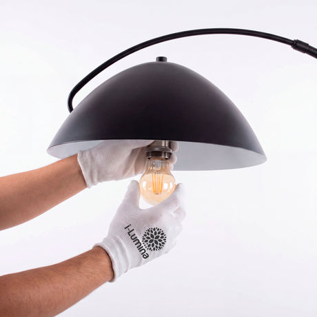 Técnico instalando bombilla de filamento en pantalla semiesférica metálica negra con guantes de protección.