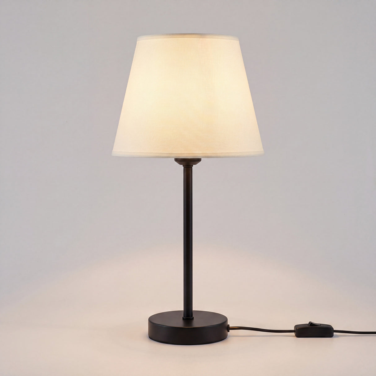 Lámpara de mesa NORA encendida con luz cálida sobre fondo neutro