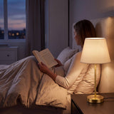Lámpara de velador NORA GOLD encendida iluminando la lectura nocturna en la cama