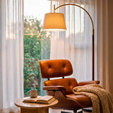 Sillón de cuero clásico en rincón de lectura iluminado por lámpara de pie de arco dorada.